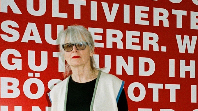 Die Galeristin Monika Sprüth steht vor einer roten Wand mit weißer Schrift.&nbsp;