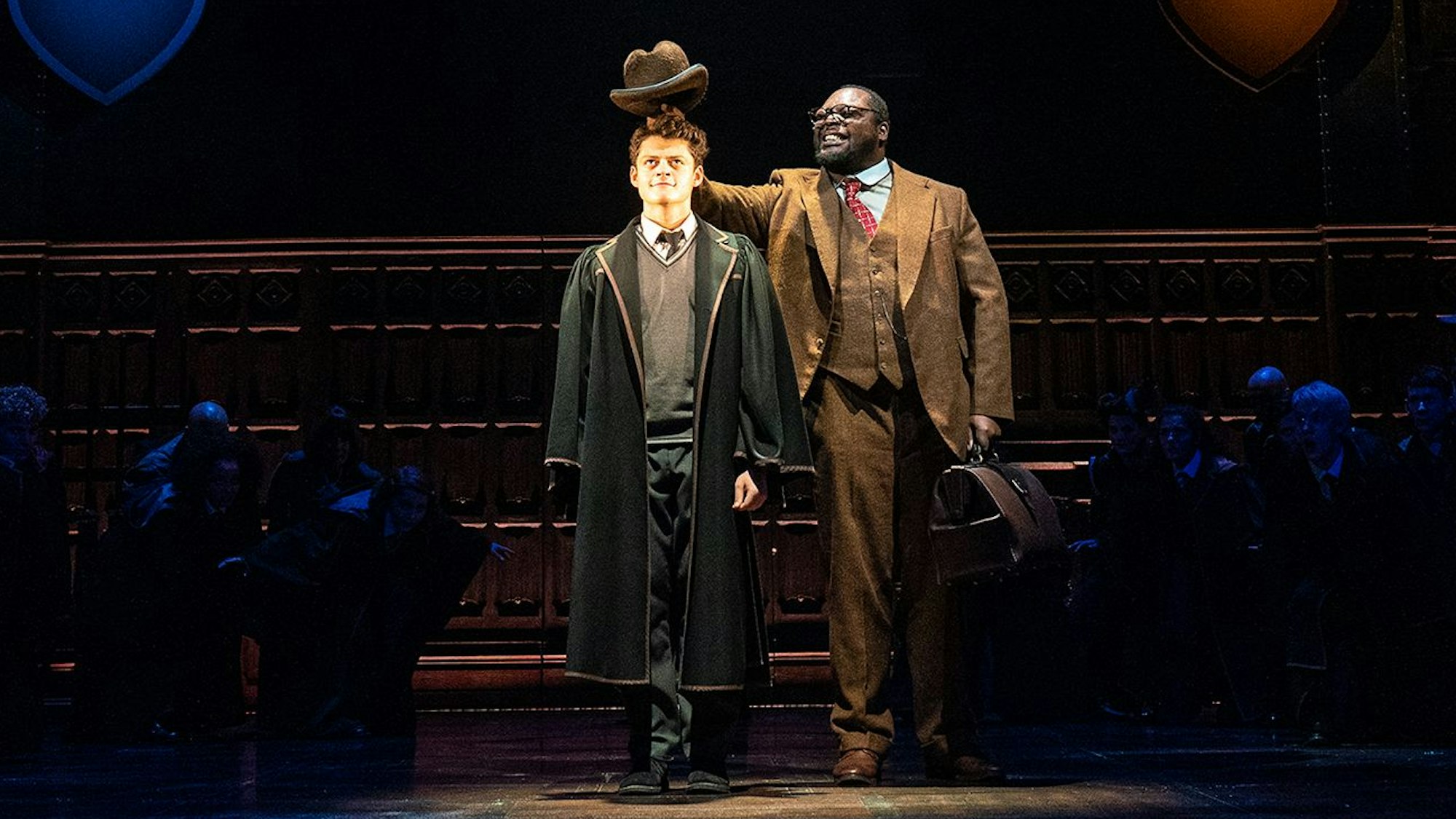 Albus Dumbledore Potter und ein Schauspieler, der den sprechenden Hut verkörpert, stehen nebeneinander auf der Bühne. Der Mann hält seinen Hut über den Kopf von Albus. Um sie herum sitzen im Dunklen weitere Hogwarts-Schülerinnen und Schüler.