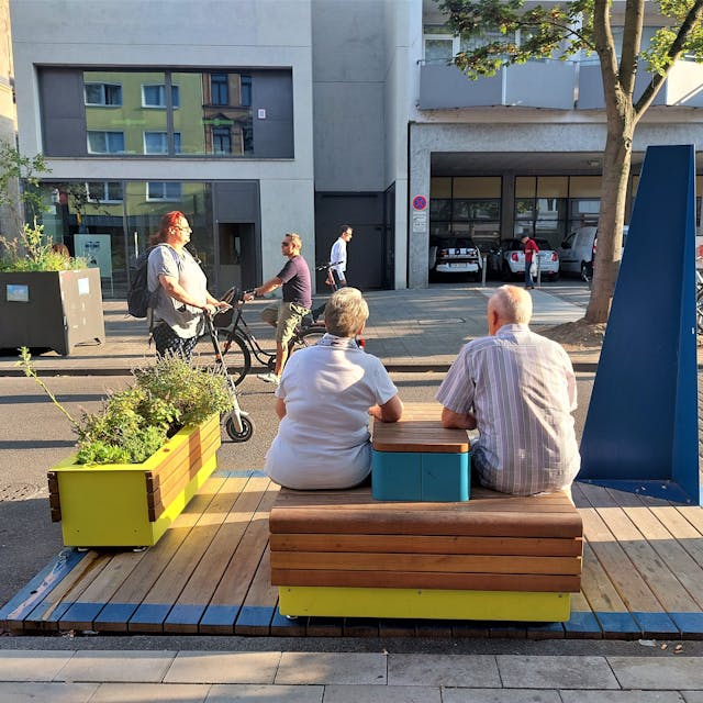 Zwei ältere Menschen sitzen auf einer Holzbank an der Deutzer Freiheit.