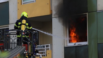 Ein Feuerwehrmann steht auf einer Drehleiter und löscht mit einem Schlauch das Feuer hinter einem Fenster. Es steigt schwarzer Rauch auf.