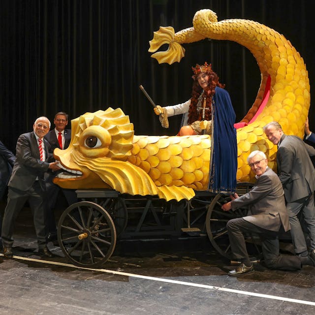 Übergabe des  historischen Festwagens an den „Held Carneval“ (Stefan F. Robert). Sechs Männer stehen um einen kleineren Karnevalswagen herum. Auf dem Wagen sitzt der „Held Carneval“ auf einem gelben Wasser-Fabelwesen.


