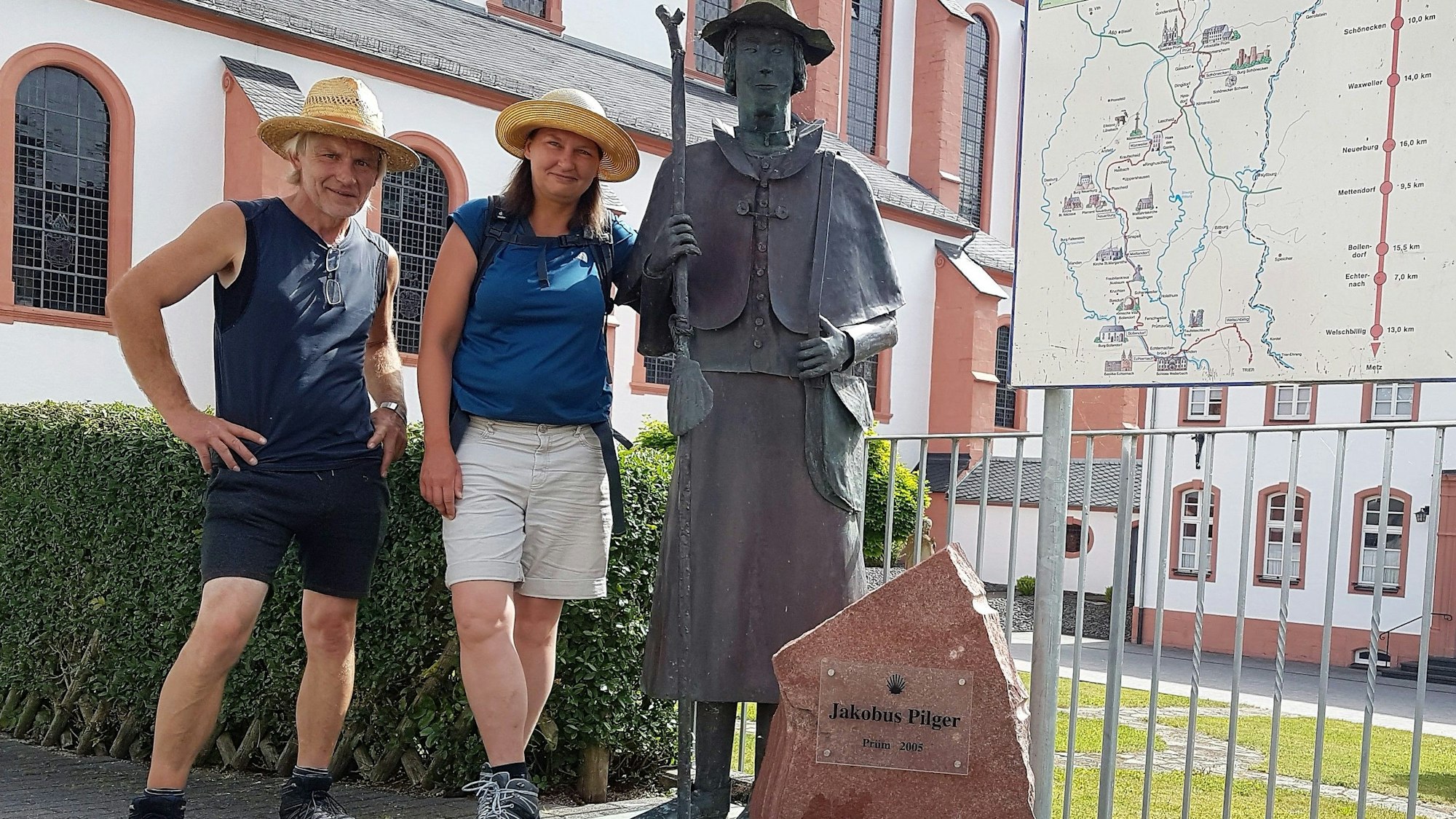 Katrin Kraemer und Uwe Sawade stehen in kurzer Hose, T-Shirt und Hut bekleidet vor einer Statur von Jakobus Pilger.