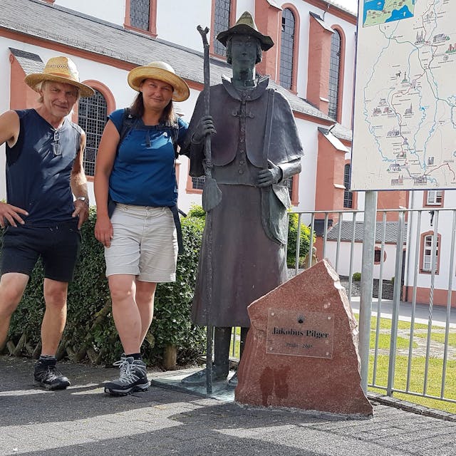 Katrin Kraemer und Uwe Sawade stehen in kurzer Hose, T-Shirt und Hut bekleidet vor einer Statur von Jakobus Pilger.&nbsp;