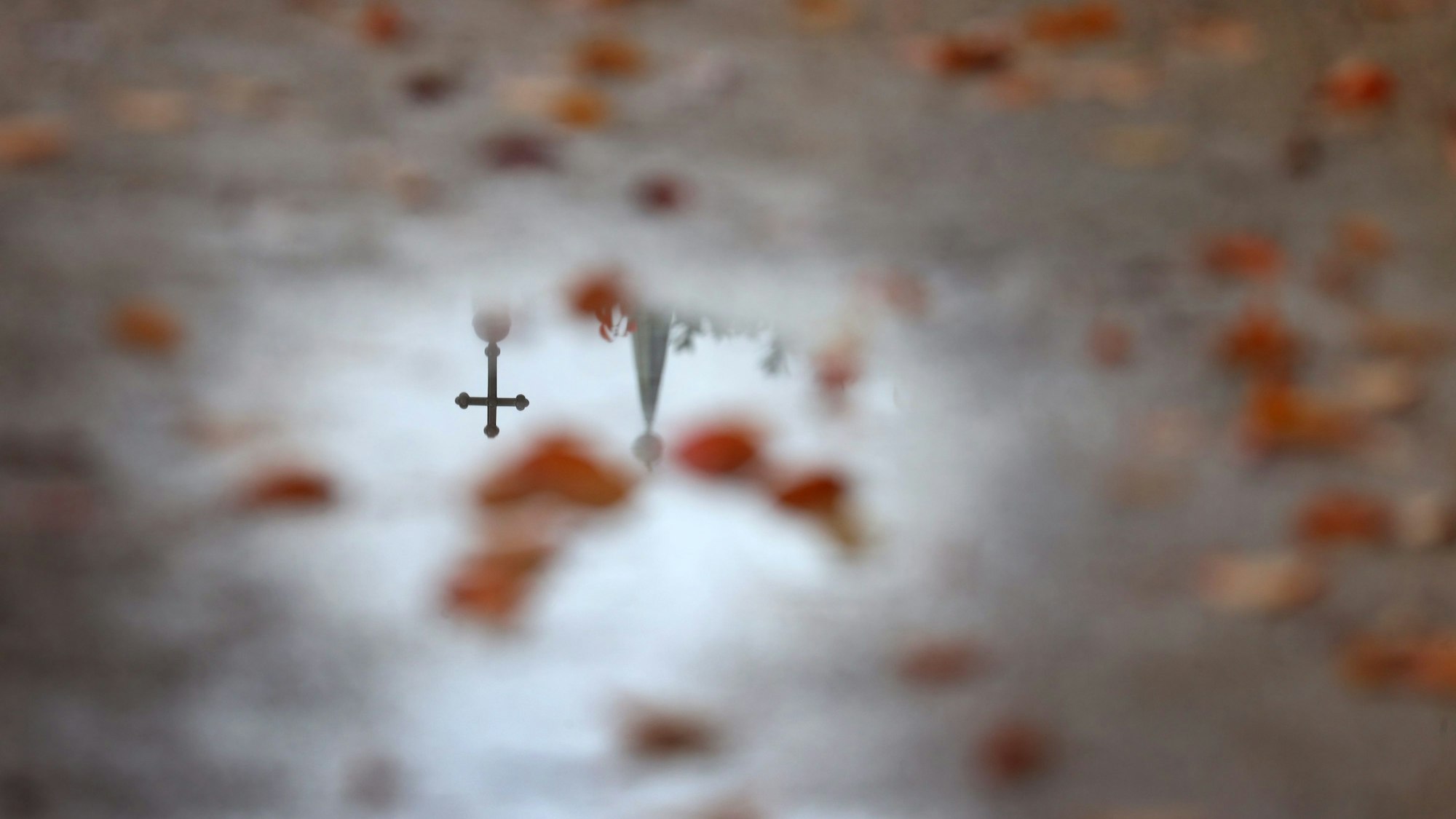 Ein Kreuz und eine Kirchturmspitze spiegelt sich in einer Wasserlache.