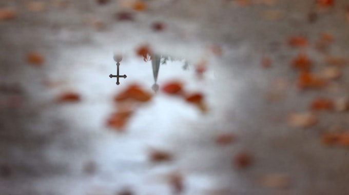 Ein Kreuz und eine Kirchturmspitze spiegelt sich in einer Wasserlache.