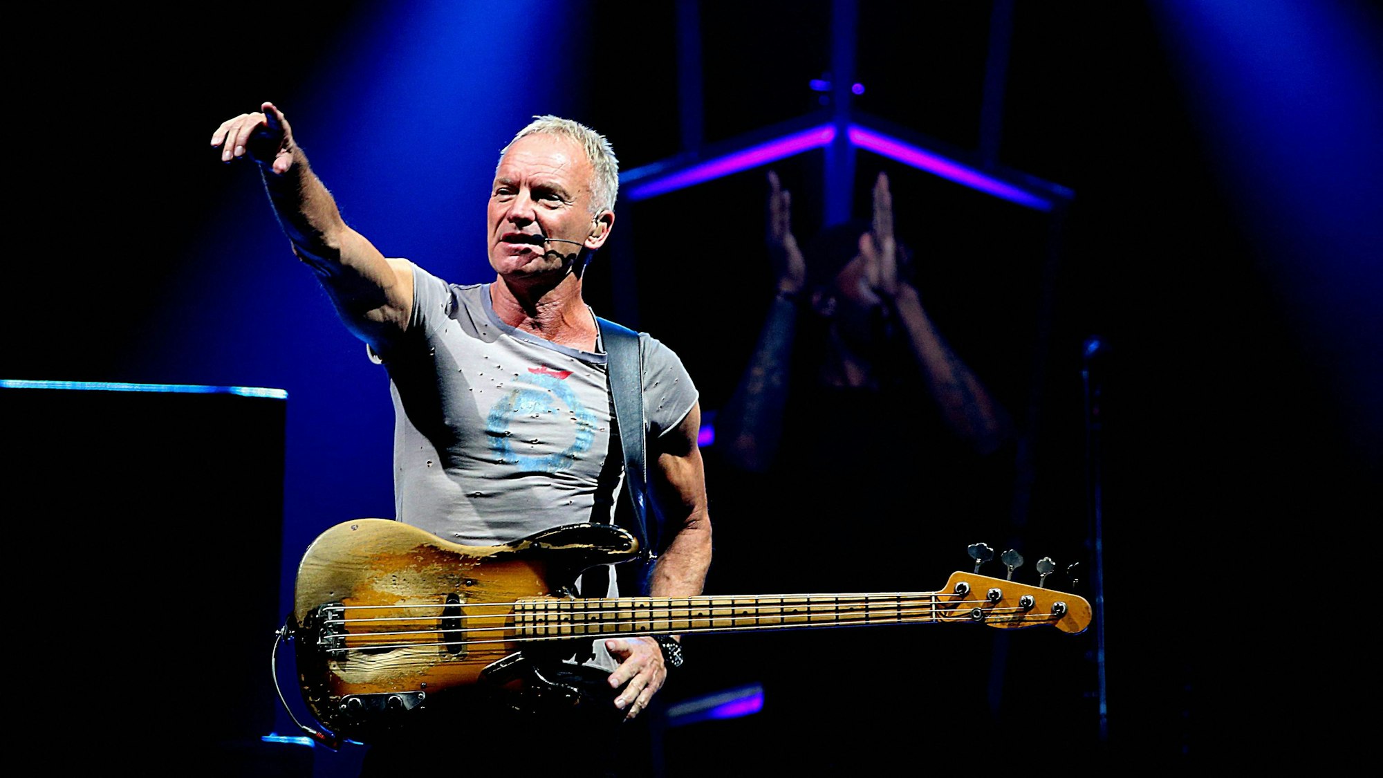 Sting steht im Rampenlicht, trägt ein graues T-Shirt und hat seine E-Gitarre umgehangen.