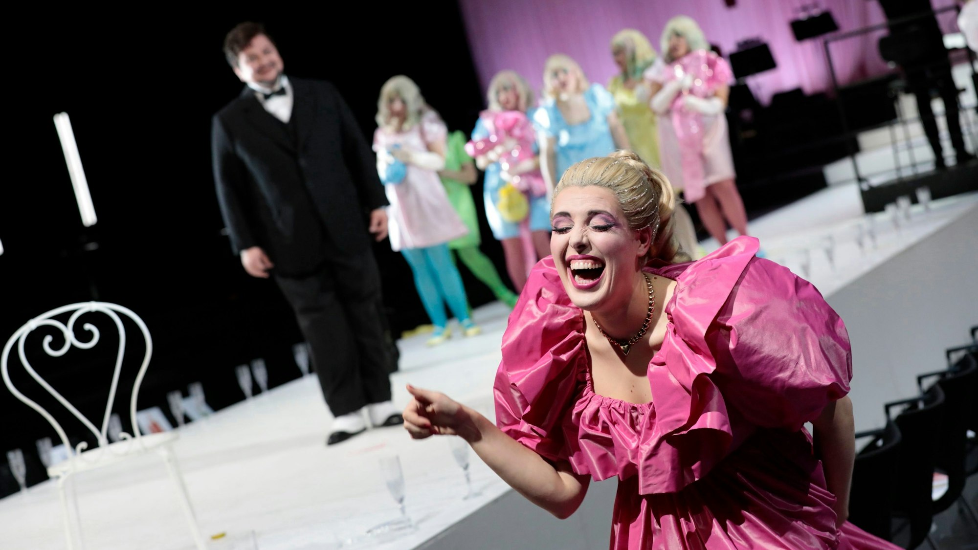 Eine Frau im rosa Rüschenkleid lacht schallen, im Hintergrund schaut die Titelfigur der Oper etwas ratlos drein.
