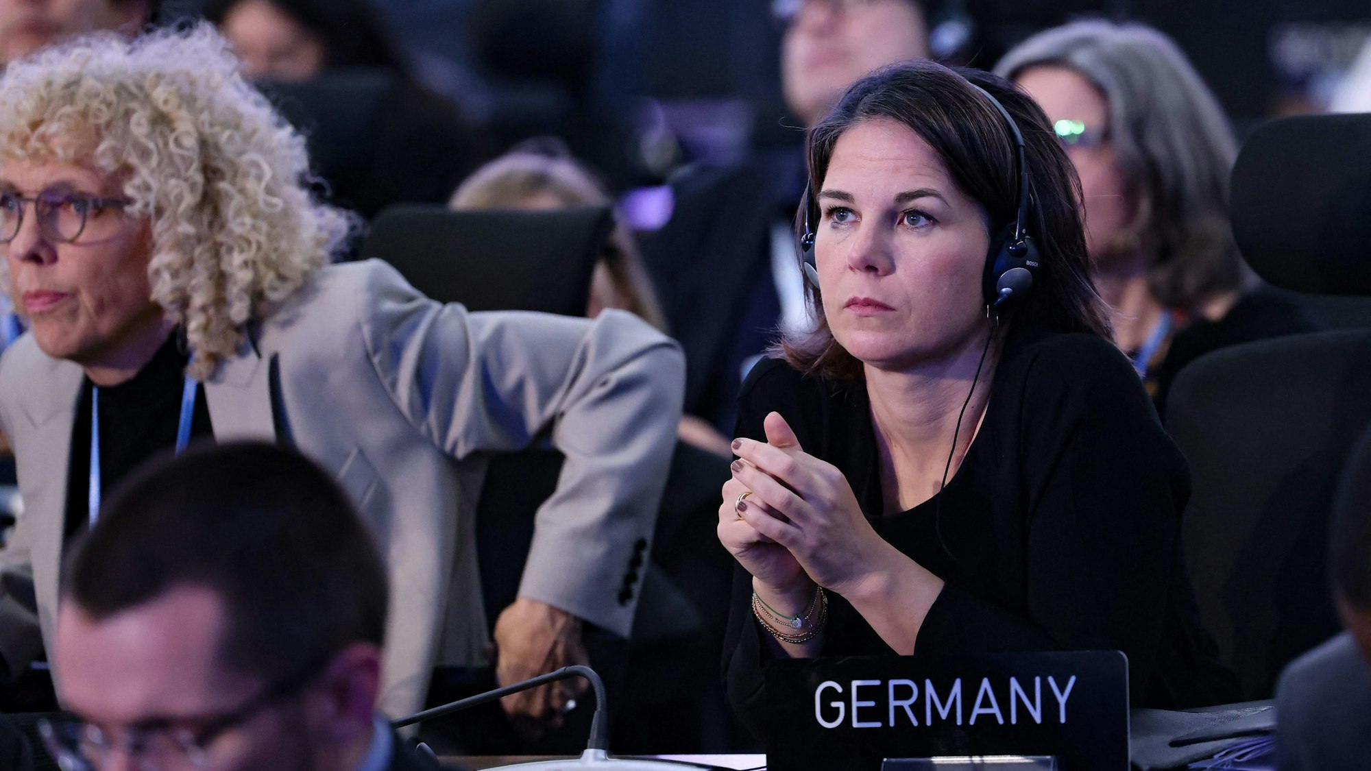Bundesaußenministerin Annalena Baerbock (Grüne) schaut skeptisch und hat in einem Konferenzsaal Kopfhörer auf. Vor ihr steht ein Schild, auf dem „Germany“ steht.