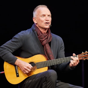 Zu sehen ist Sting, wie er auf der Bühne sitzt, er hält eine Gitarre in den Händen und singt.