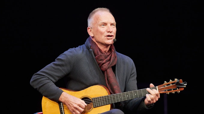 Zu sehen ist Sting, wie er auf der Bühne sitzt, er hält eine Gitarre in den Händen und singt.