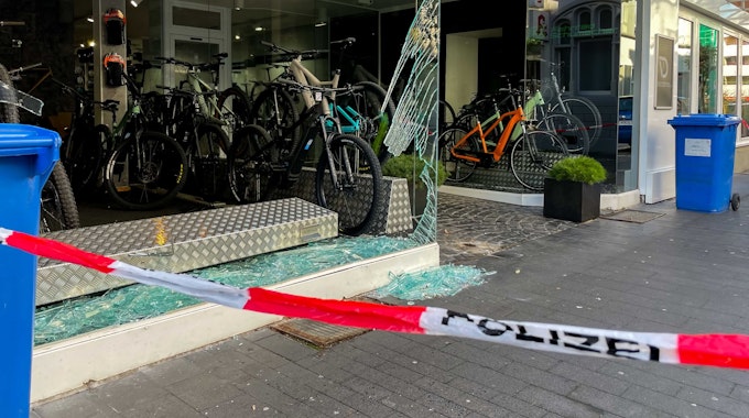 Das zertrümmerte Schaufenster eines Fahrradladens. Auf dem Boden und in der Auslage liegen Glasscherben, einige Fahrräder sind noch ausgestellt. Im Vordergrund steht auf weiß-rotem Absperrband das Wort Polizei.