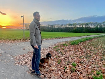 Ein Mann blickt auf ein Feld, sein Hund sitzt neben ihm.