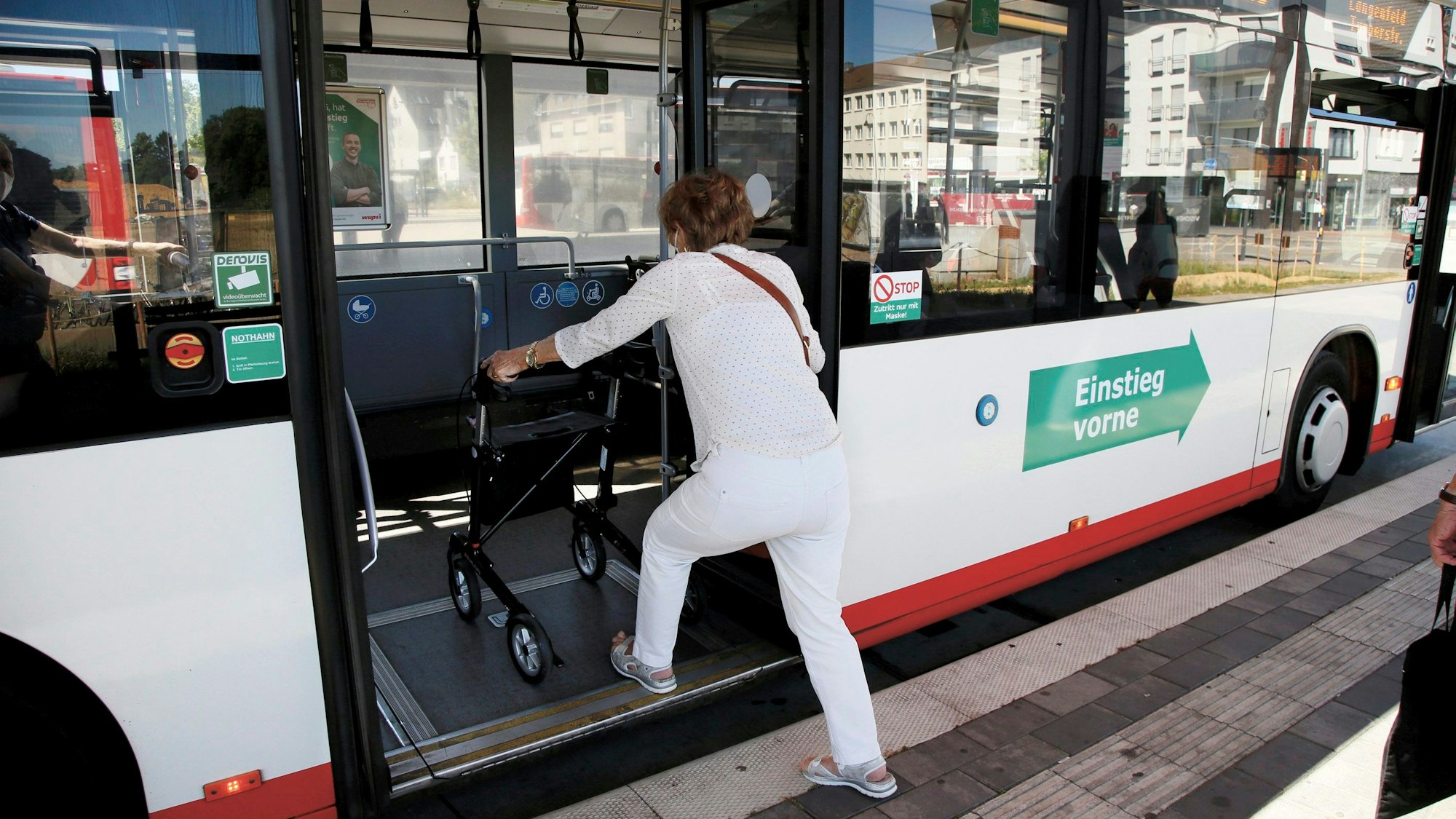 Eine ältere, weiß gekleidete Frau steigt in die hintere Tür eines Linienbusses ein. Sie schiebt einen Rollator.