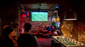 Im „Stiefel“ auf der Zülpicher Straße verfolgten einige wenige Kölner das WM-Auftaktspiel der deutschen Mannschaft gegen Japan.Das Bild zeigt drei junge Männer an einem Tisch, im Hintergrund hängt der TV-Bildschirm.