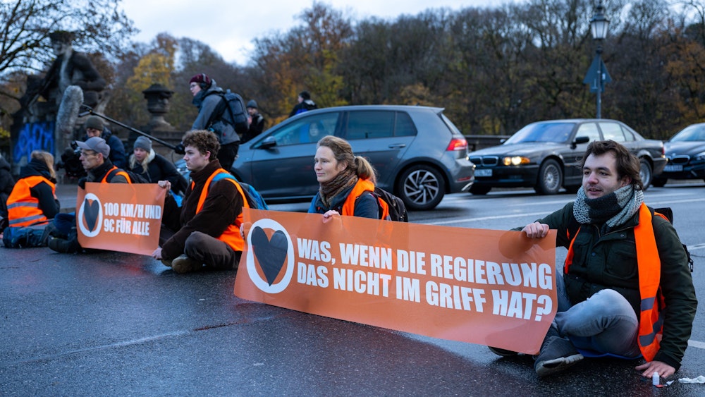 Klimaaktivisten der Umweltschutzbewegung „Letzte Generation“ sitzen auf der Prinzregentenstraße in München und blockieren den Verkehr. Sie halten Banner mit dn Aufschriften „Was, wenn die Regierung das nicht im Griff hat“ und „100 km/h und 9 € für alle“