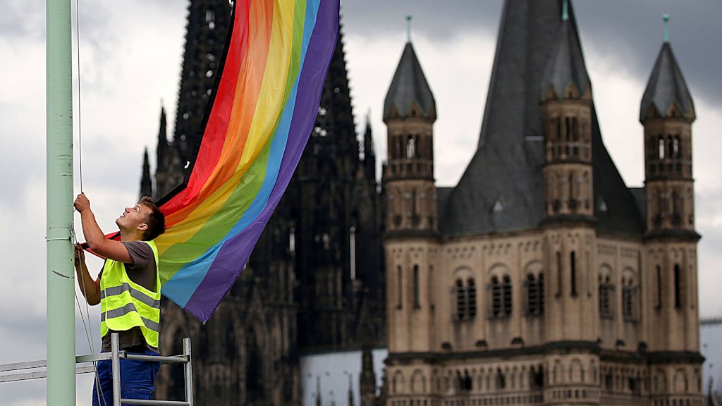 Ein Arbeiter hängt in Köln vor der Kulisse des Doms und der Kirche Groß St. Martin eine Regenbogenfahne auf.&nbsp;
