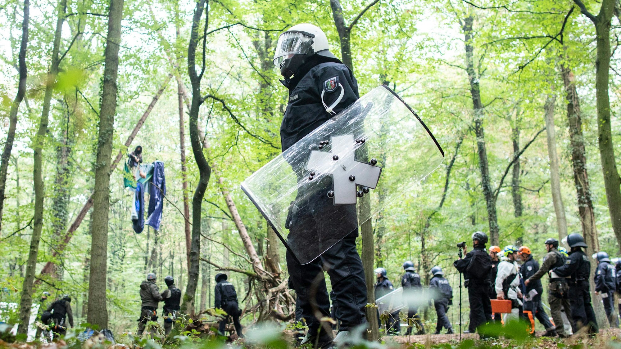 Polizisten stehen im Wald vor einer Barrikade, in der sich eine Aktivistin festgebunden hat. Vorne im Bild ist ein Polizist mit Schild und Helm zu sehen.