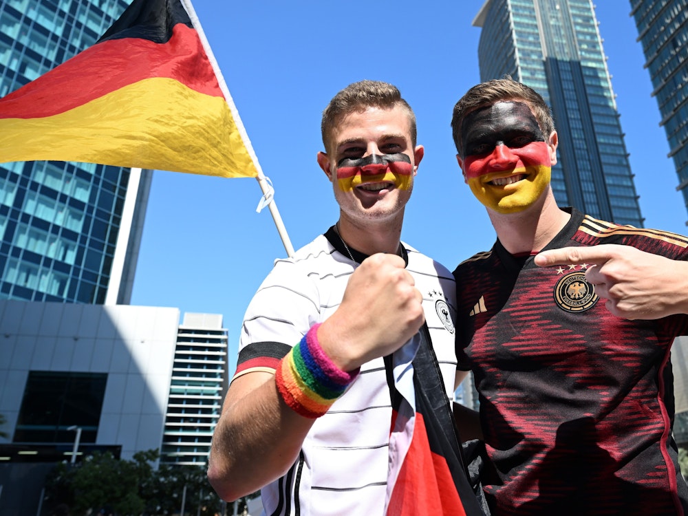 Bengt Kunkel hält sein Armband in Regenbogenfarben in die Kamera, neben ihm steht sein Bruder Jan. Beide haben ihre Gesichter in den Deutschland-Farben bemalt.