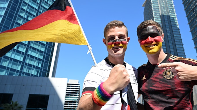 Bengt Kunkel hält sein Armband in Regenbogenfarben in die Kamera, neben ihm steht sein Bruder Jan. Beide haben ihre Gesichter in den Deutschland-Farben bemalt.
