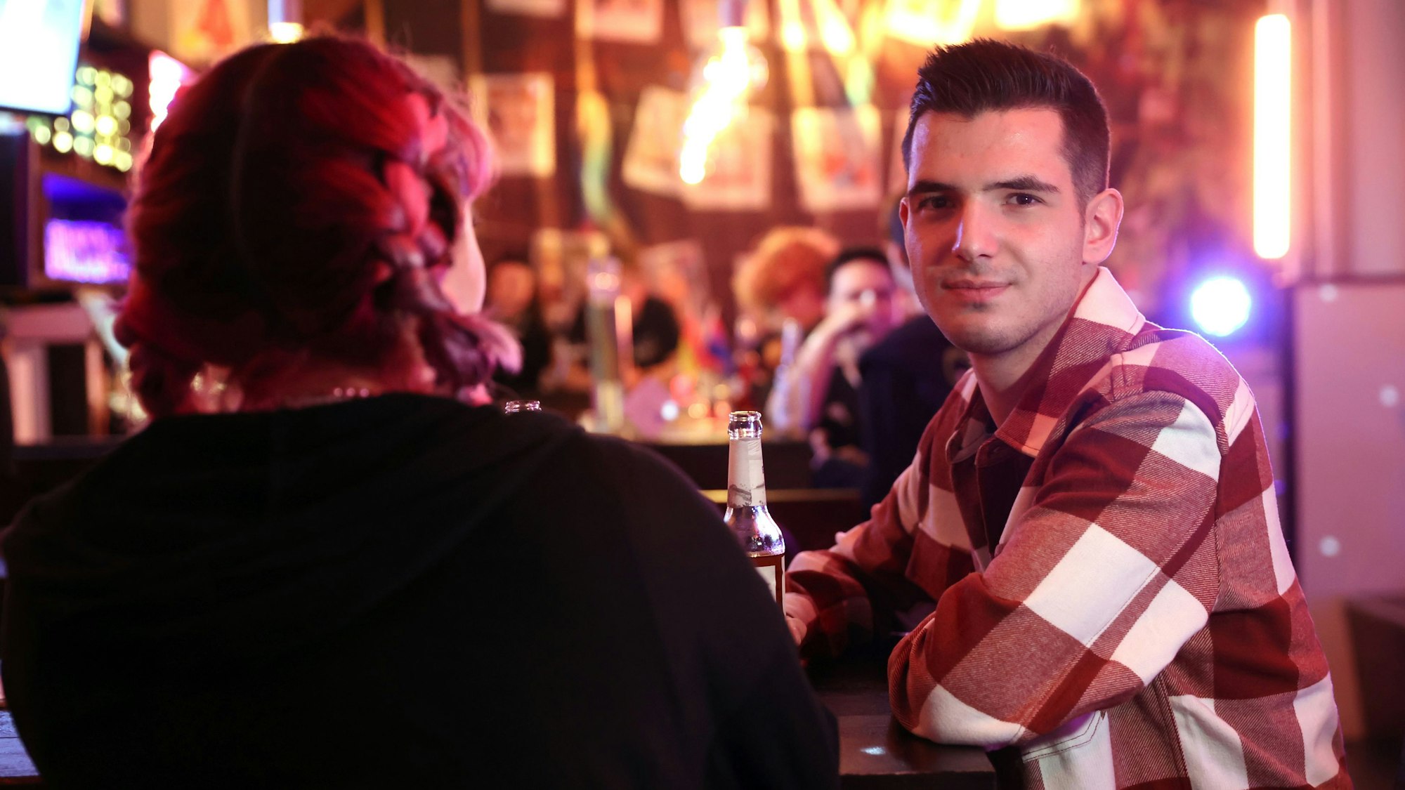 Zwei queere Jugendliche an der Bar des Kölner Jugendzentrums „Anyway“. Links mit dem Rücken zur Kamera sitzt Kitti, mit pinken Zöpfen. Daneben sitzt David im karierten Hemd und schaut in die Kamera.
