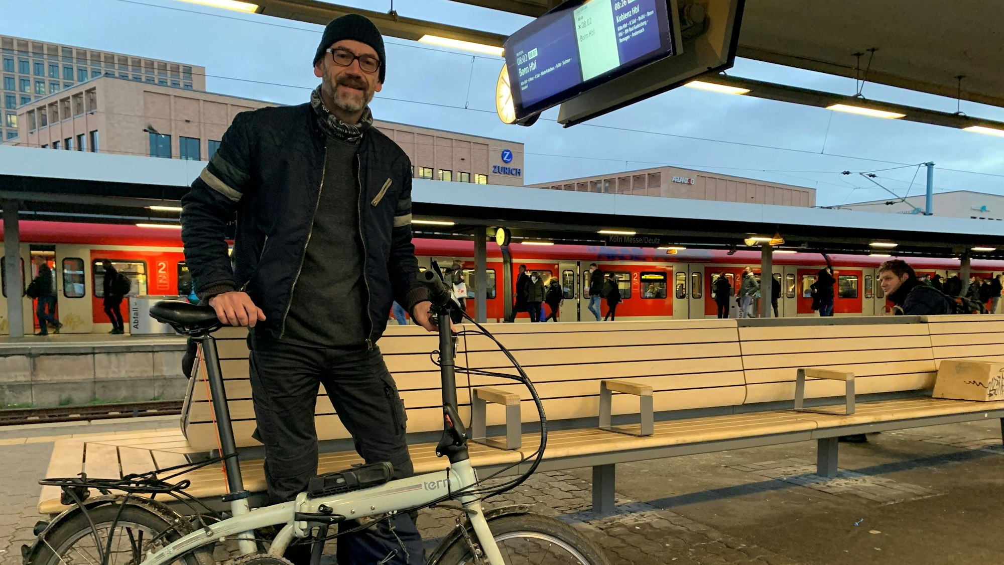 Bahnpendler Michael Weber steht mit seinem Faltrad am Bahnhof Köln-Messe/Deutz und wartet auf seinen Zug Richtung Pulheim. Im Hintergrund ist eine S-Bahn auf dem Gleis zu sehen.