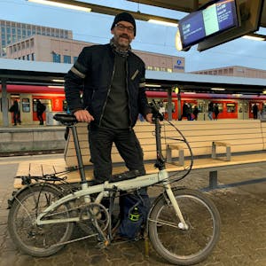 Bahnpendler Michael Weber steht mit seinem Faltrad am Bahnhof Köln-Messe/Deutz und wartet auf seinen Zug Richtung Pulheim. Im Hintergrund ist eine S-Bahn auf dem Gleis zu sehen.