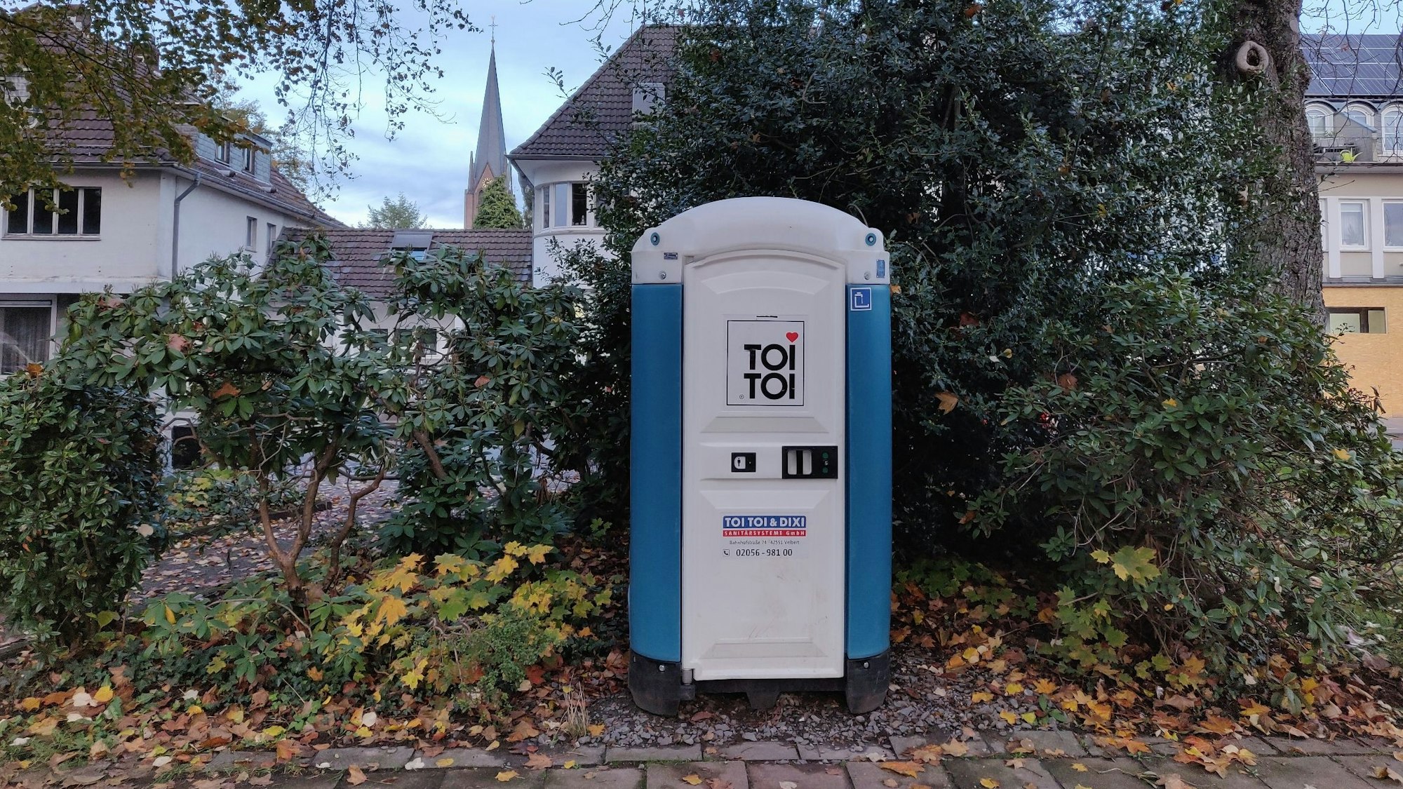 Eine blau-weiße Mobiltoilette steht unter Bäumen im Stadtpark am Busbahnhof Leichlingen