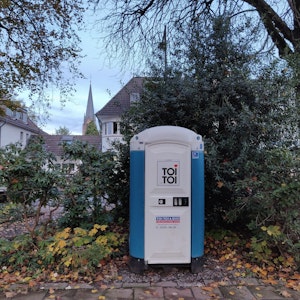 Eine blau-weiße Mobiltoilette steht unter Bäumen im Stadtpark am Busbahnhof Leichlingen