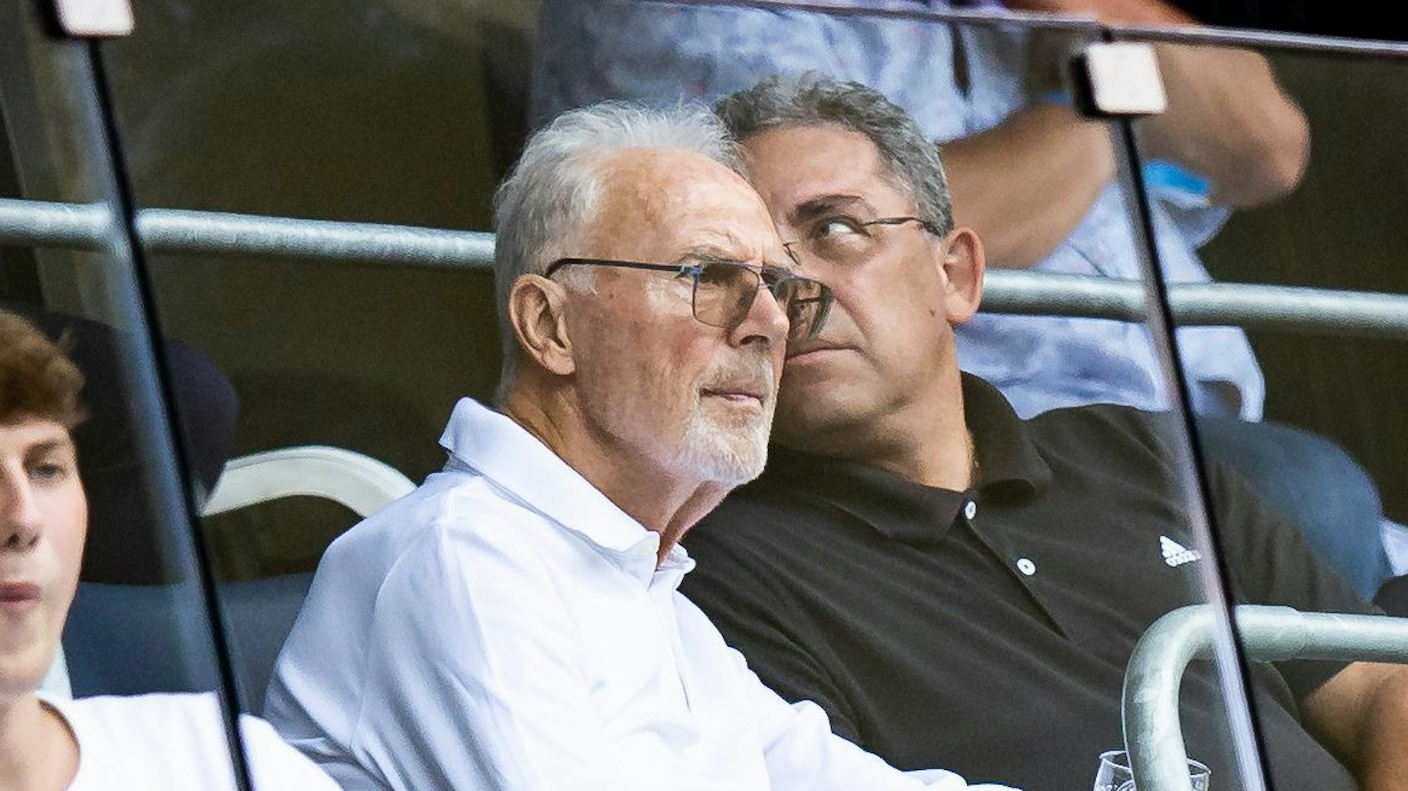 Franz Beckenbauer sitzt im Publikum bei einem Fußballspiel. Neben ihm ein Mann. Beckenbauer trägt eine getönte Brille und schaut mit ernster Miene auf das Spielfeld. Er trägt ein weißes Oberteil.