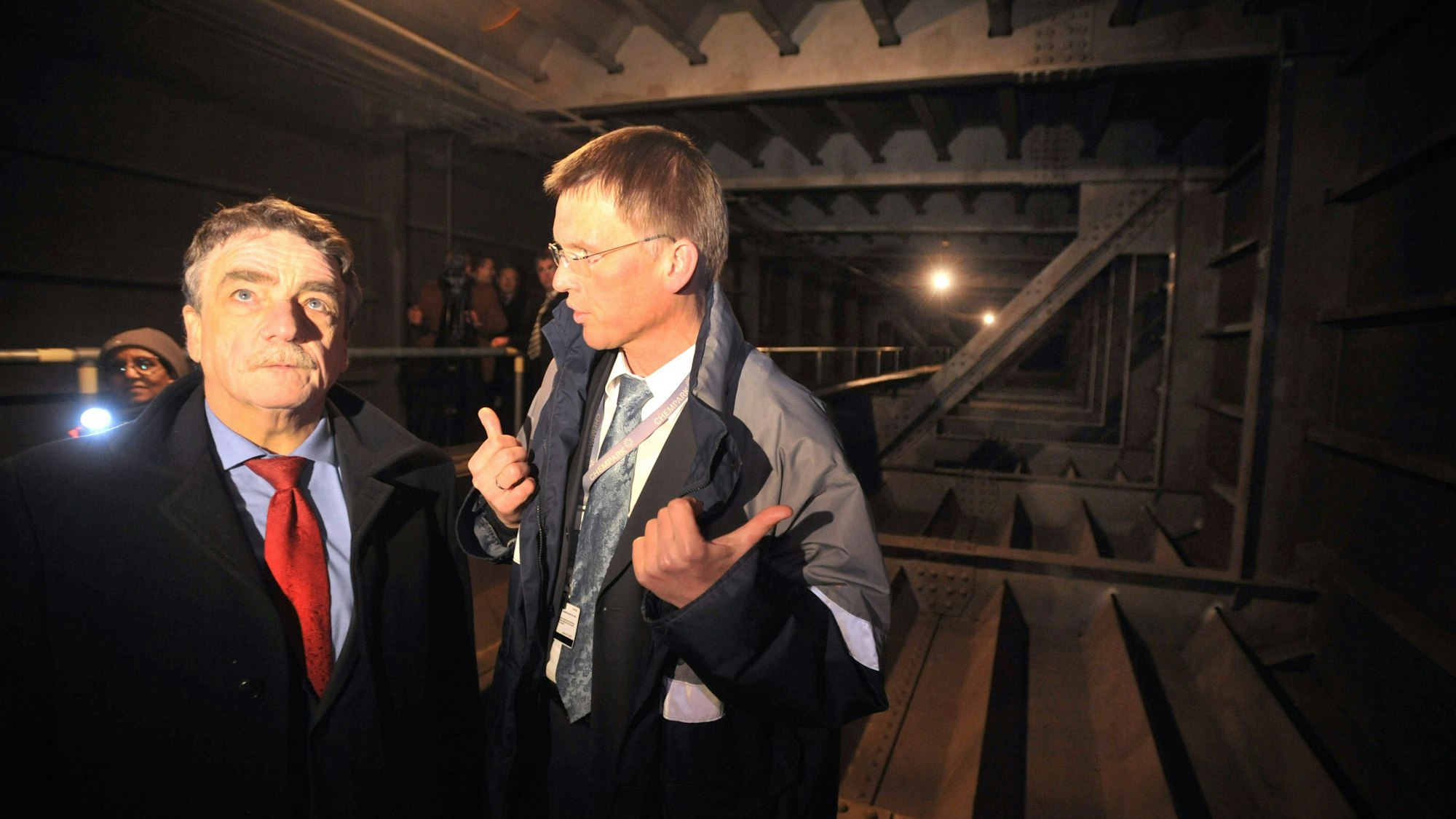NRW-Verkehrsminister Groschek und Ernst Grigat, Leiter Chemparks Leverkusen stehen im Brückenkasten der Leverkusener Brücke. Es ist eine dunkle Stahlkonstruktion. Im Hintergrund leuchtet jemand mit einer Taschenlampe.
