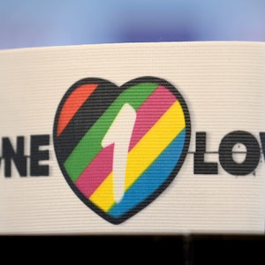 Eine Armbinde, die ein Herz und die Aufschrift „One Love“ trägt, steht auf einer Oberfläche. Das Herz ist mit bunten Streifen sowie der Zahl 1 gefüllt. Die Farben symbolisieren die Vielfalt unterschiedlicher Lebensentwürfe.