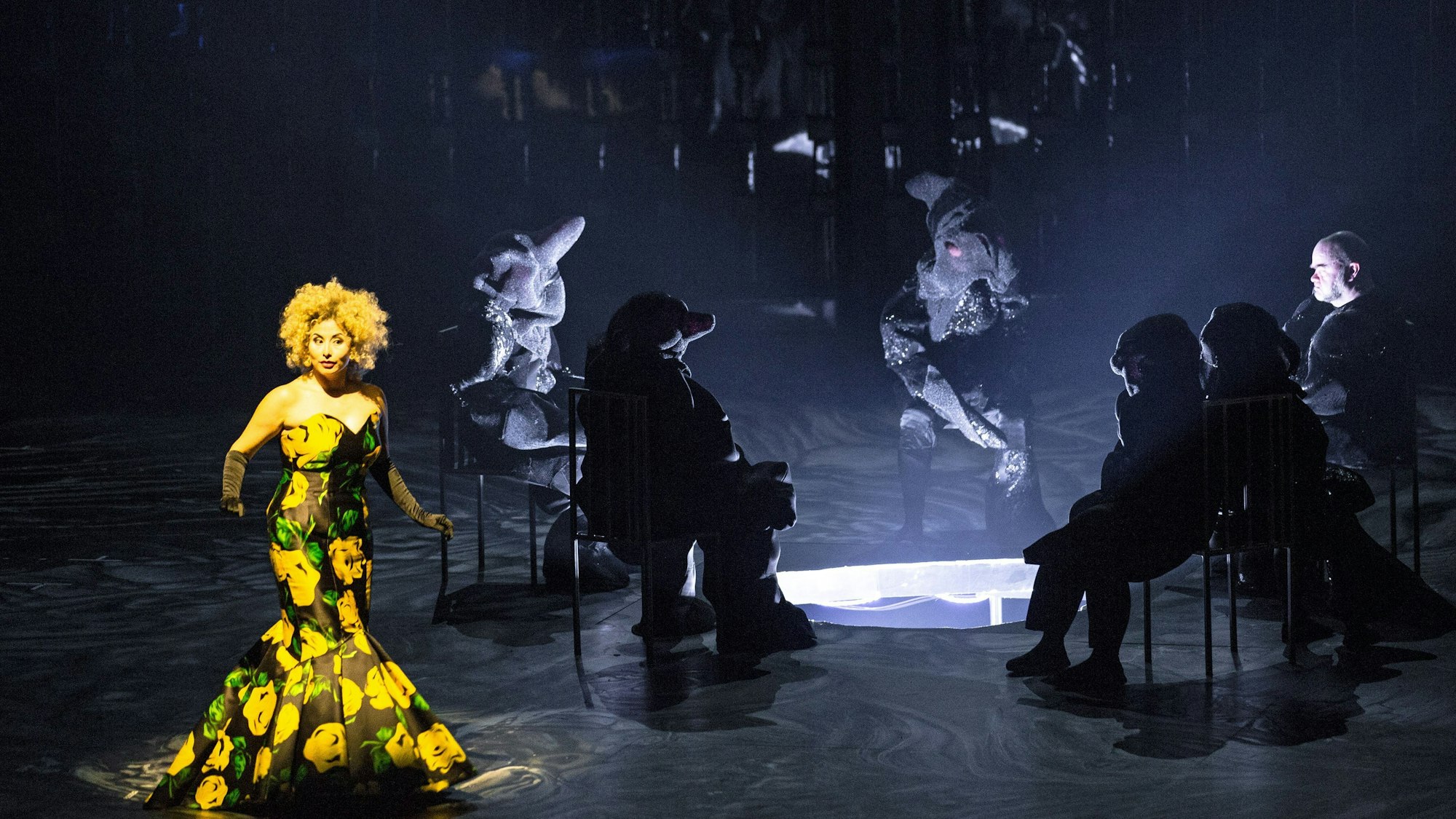 Oper Köln DER MEISTER UND MARGARITA Musik. Eine gelb angestrahlte Frau in einem Kleid geht über die Bühne, hinter ihr sitzen dunkle und graue Gestalten auf Stühlen im Kreis.