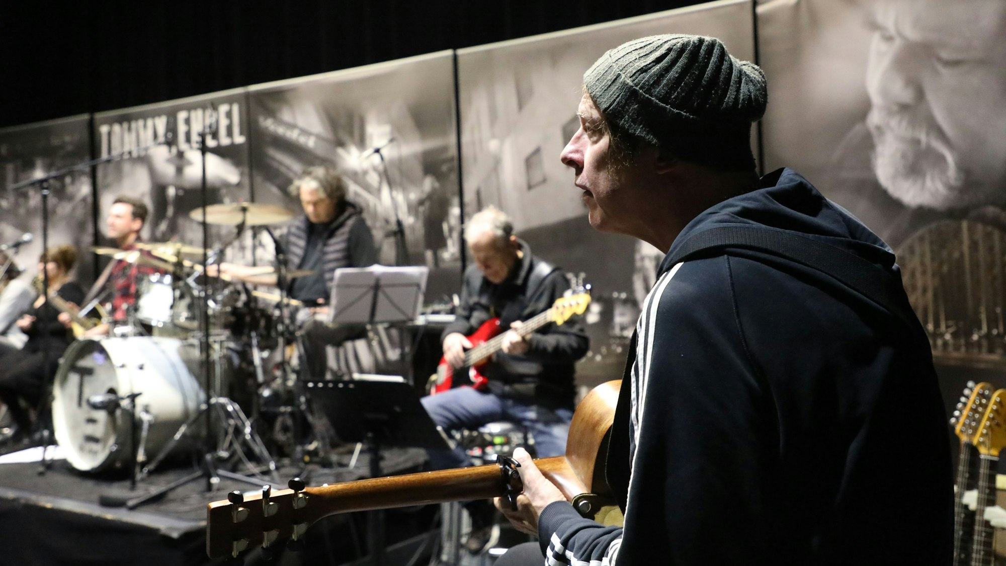 Ein Mann mit Mütze hält eine Gitarre in der Hand, im Hintergrund sind ein Bassist, ein Schlagzeuger und einer weiterer Gitarrist zu sehen.