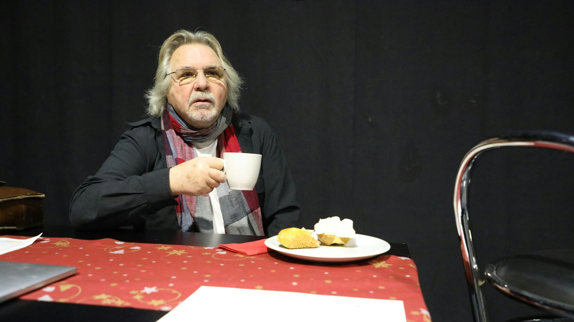 Der Sänger Tommy Engel sitz mit einer Tasse Kaffee und einem Käsebrötchen an einem Tisch mit Weihnachtstischdecke.
