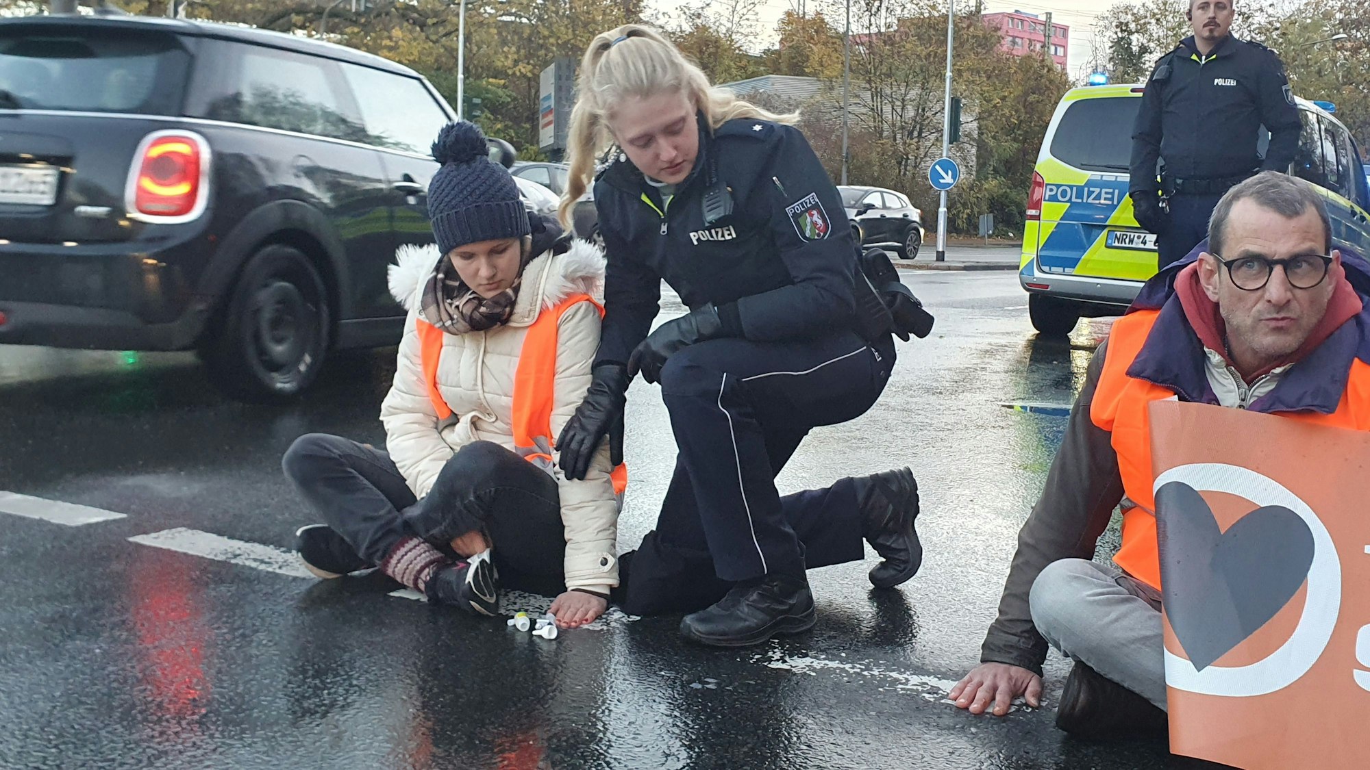 Eine Polizistin kniet auf einer Straße neben zwei Klimaschutzaktivisten.