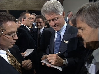 Das Bild zeigt den ehemaligen US-Präsidenten Bill Clinton. Mit einem Stift schreibt er etwas auf einen Zettel. Um ihn herum stehen weitere Männer.