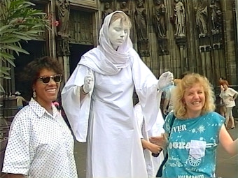 Das Bild zeigt eine junge Frau in der Mitte, die ganz in Weiß gekleidet ist und das Gesicht weiß bemalt hat. Sie hat die Augen geschlossen. Neben ihr stehen zwei lachende Frauen.