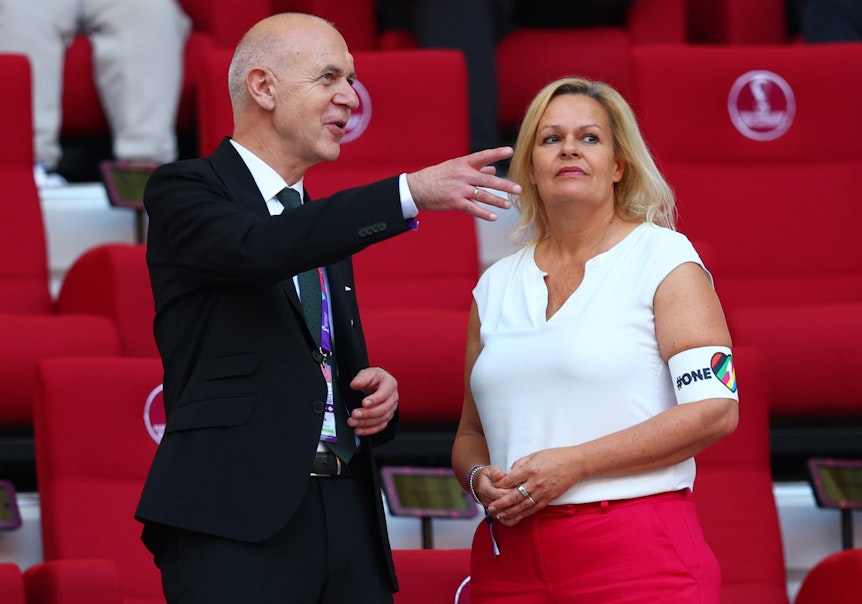 Bundesinnenministerin Nancy Faeser (SPD) und DFB-Präsident Bernd Neuendorf unterhalten sich im Stadion in Katar. Frau Faeser trägt ein ärmelloses weißes Shirt und an ihrem Oberarm eine Armbinde mit der Aufschrift „One Love“.