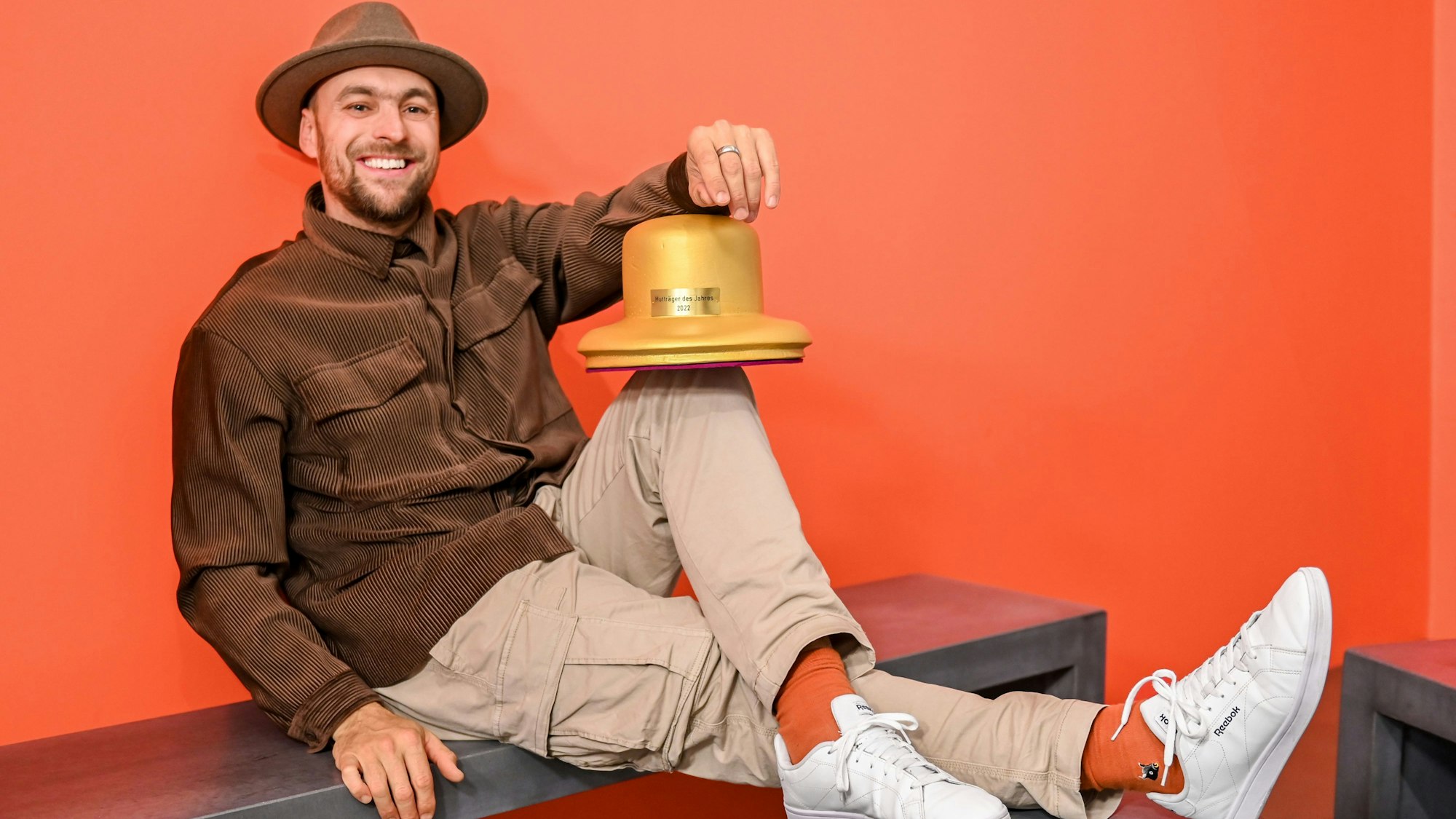 Der Musiker Max Mutzke posiert mit dem Goldenen Hut-Award und sitzt auf einer Bank in einem Raum mit roten Wänden.