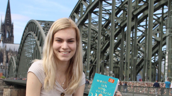 Antonia Wesseling sitzt am Rhein auf einer Mauer, hinter ihr die Hohenzollernbrücke und der Dom. In den Händen hält sie ihren Roman "Wenn ich uns verliere".