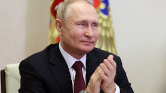 Wladimir Putin sitzt in seinem Büro am Schreibtisch und lächelt.