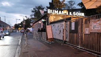 Außenaufnahmen vor dem Bumann und Sohn, links fahren Autos, rechts oben ist der leuchtende Schriftzug der Bar zu sehen, davor stehen Menschen an, um in den Laden zu kommen.