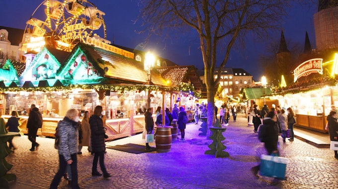 Die Weihnachtsmarktbuden leuchten im Dunkeln. Menschen gehen einzeln oder in kleinen Gruppen zwischen den Buden.