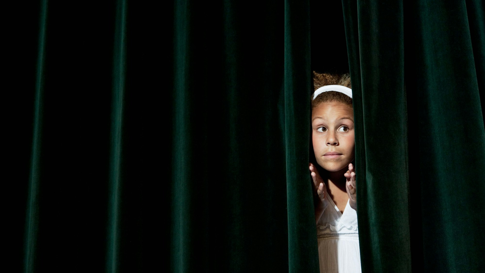 Ein junges Mädchen schaut verschüchtert durch einen grünen Samtvorhang.
