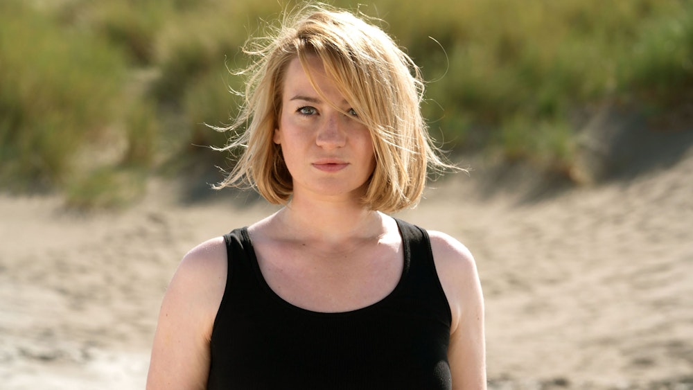 Nina Gummich in ihrer Rolle Alice Schwarzer, sie steht am Strand, hat ein schwarzes Top an