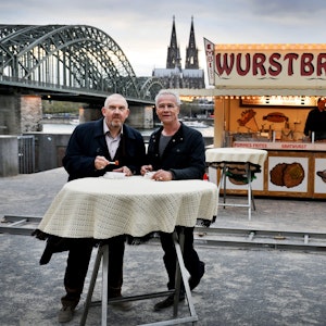 Die beiden Kölner Tatort-Kommissare stehen vor einer Bratwurst-Bude und dem Dom-Panorama.