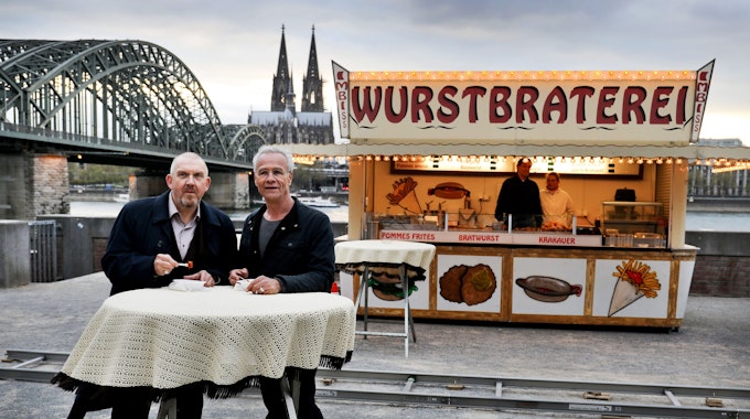 Die beiden Kölner Tatort-Kommissare stehen vor einer Bratwurst-Bude und dem Dom-Panorama.