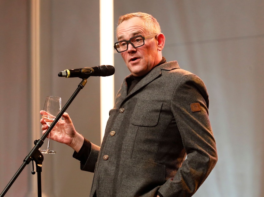 Mann in grauem Anzug steht entspannt mit einem Glas Wein vor einem Mikrofon und spricht mit dem Publikum.