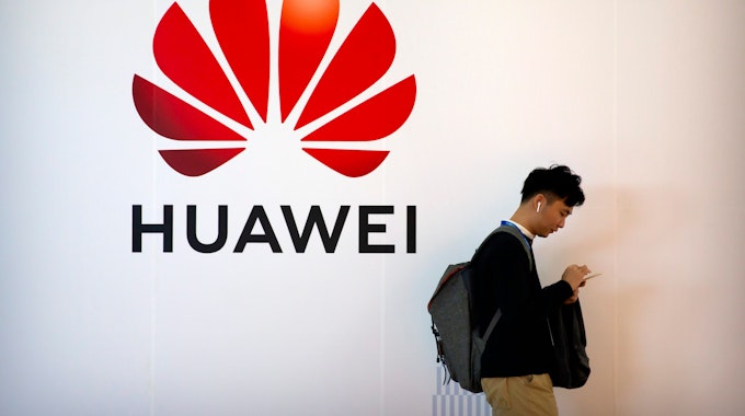 Ein Mann tippt auf seinem Smartphone, während er auf der Fachmesse PT Expo in der Nähe einer Werbetafel für die chinesische Technologiefirma Huawei steht.