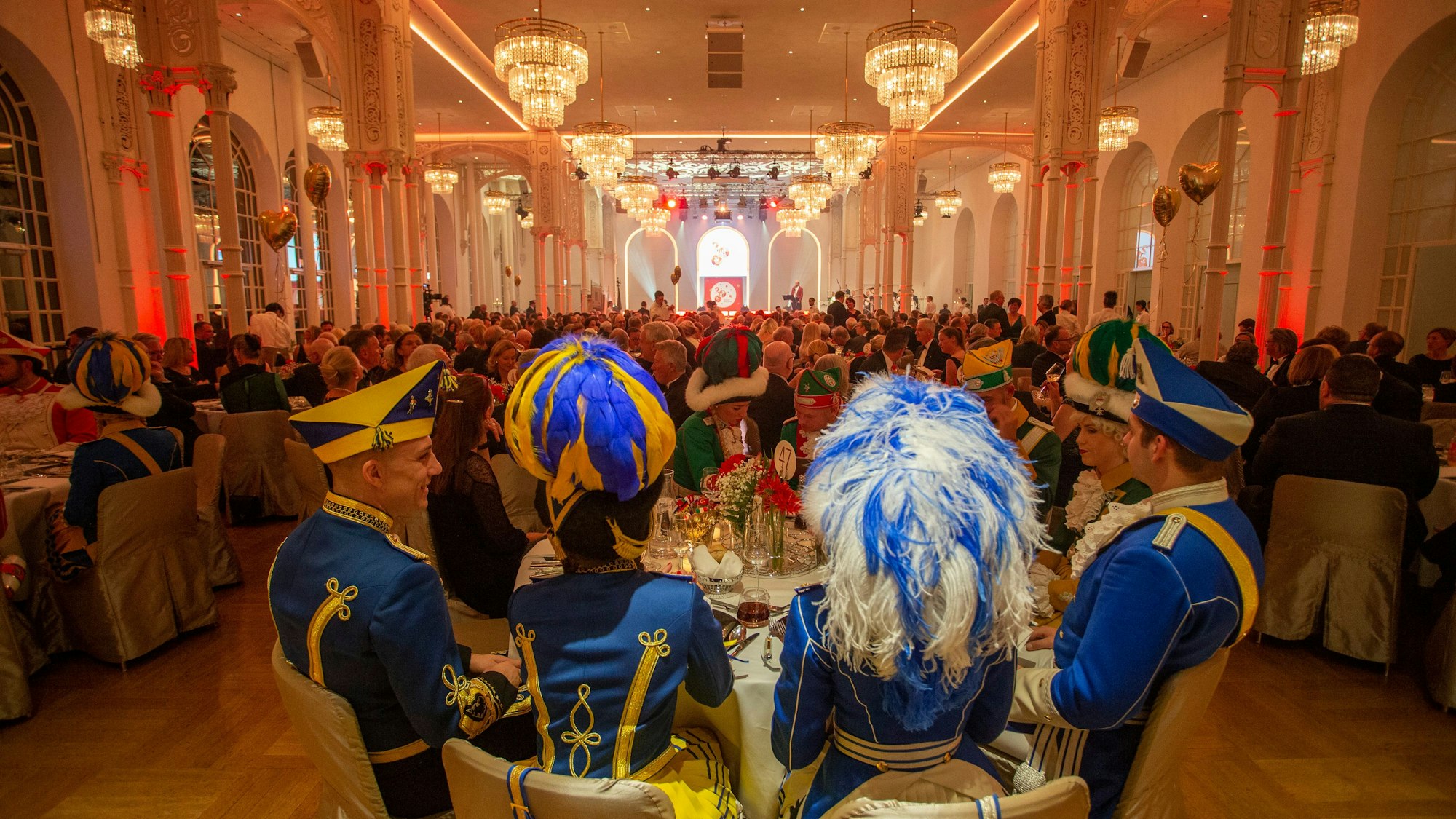 In der prunkvoll gestalteten Flora sitzen rund 500 Gäste an runden Tischen. Im Vordergrund sitzen Mitglieder der Tanzgruppen des Kölner Traditionskorps.
